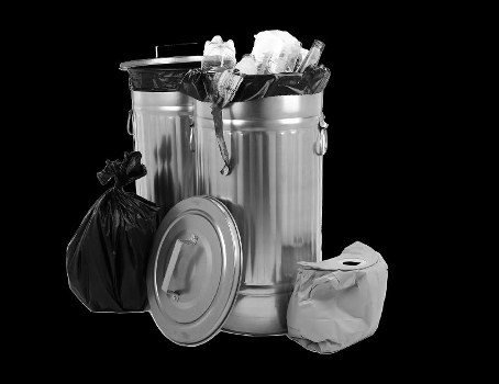A large metallic waste bin filled with crumpled paper, plastic bottles, and other rubbish, situated on a dark background. The bin has a lid leaning against its side and is lined with a partially open black plastic bag containing additional waste. To the left of the bin, there is a tightly tied black plastic rubbish bag, and to the right, a crumpled paper bag is placed on a flat surface. The scene appears to be a controlled environment, possibly a designated rubbish collection point or a cleaning area. The metallic surface of the bin reflects minimal light, highlighting its cylindrical shape and polished finish. The overall composition emphasizes the collection of general waste, consistent with a professional rubbish removal service such as Commercial Waste Removal Sanderstead, operating within the local Sanderstead area postcode context.