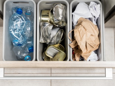 The image shows three grey plastic bins placed in a row on a tiled floor, each filled with different types of waste. The left bin contains several transparent plastic bottles, some partially crushed, with blue caps visible. The middle bin holds a collection of metallic, aluminum cans, some crushed or bent, with a reflective surface and ridged sides. The right bin contains crumpled paper and brown paper bags, with some sheets of white paper mixed in, creating a disorganized pile. The bins are situated outdoors near a building's exterior wall, and the scene is lit with natural daylight, highlighting the textures of the plastic, metal, and paper waste. This setup is representative of waste left for collection, aligned with commercial rubbish removal services like those offered by Commercial Waste Removal Sanderstead, operating in the local postcode area to Sanderstead, South London.