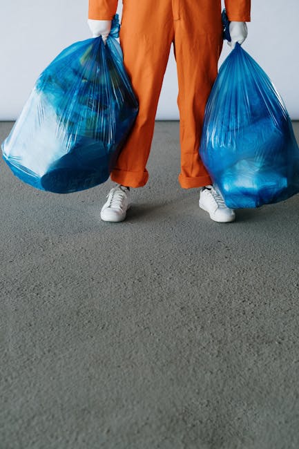A person dressed in orange overalls and white sneakers standing on a grey floor, holding two large blue plastic rubbish bags filled with waste materials, with their arms extended downward. The backgro