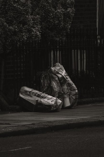 A person dressed in orange overalls and white sneakers standing on a grey floor, holding two large blue plastic rubbish bags filled with waste materials, with their arms extended downward. The backgro